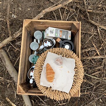 Ein kleiner Holzkasten in dem ein Berief Getränkekarton zusammen mit Tassen und Kannen, darauf ein Korb mit einem Croissant auf einem Waldboden.