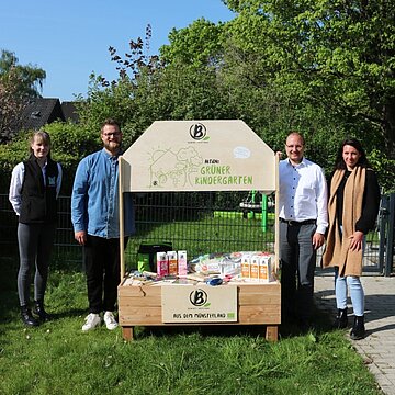 Hochbeet für die Kindertagespflege Menschen neben einem Hochbeet, auf dem Berief Getränkekartons stehen und auf den grünen Kindergarten hingewiesen wird.