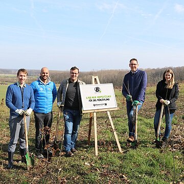 Neue Baumsetzlinge für das Münsterland Fünf Menschen stehen auf einem Feld um ein Plakataufsteller herum.