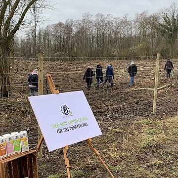 Ein Berief Plakat auf einem Aufsteller vor einem Maschendrahtzaun auf einer Brache, dahinter gehen Menschen umher.