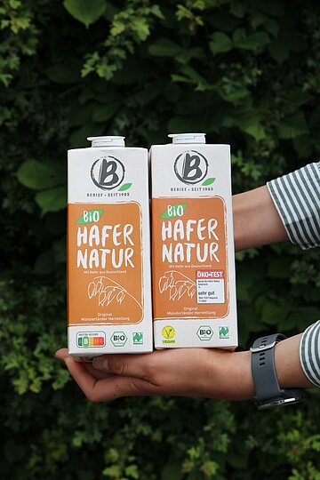 Zwei Hafergetränkekartons werden vor einem Blätterhintergrund in die Kamera gehalten.
