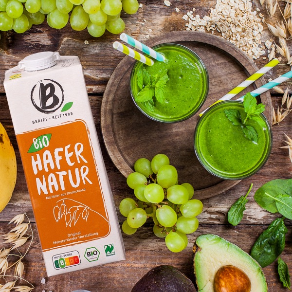 Karton Hafer-Drink neben einem Holzbrett auf dem zwei grüne Smoothies stehen mit Früchte-Dekoration