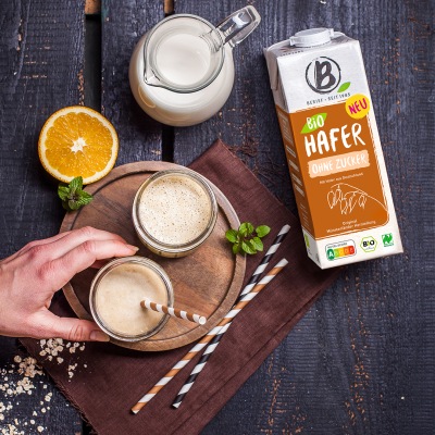 Zwei hellbraune Smoothies stehen auf einem runden Holzbrett mit Strohhalm und Dekoration