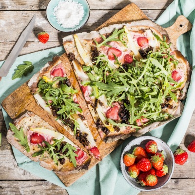 Flammkuchen belegt mit Erdbeeren, Spargel und Rucola auf einem Holzbrett
