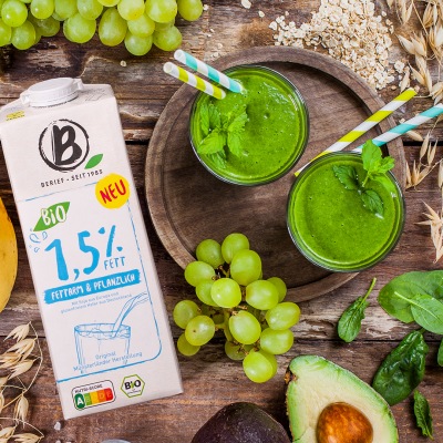 2 grüne Smoothies in Gläsern mit Strohhalm auf einem Holzbrett mit Getränkekarton.